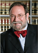 Portrait of a bespectacled man with gray beard in a black suit and red bow tie, standing in front of a law library with stacked books
